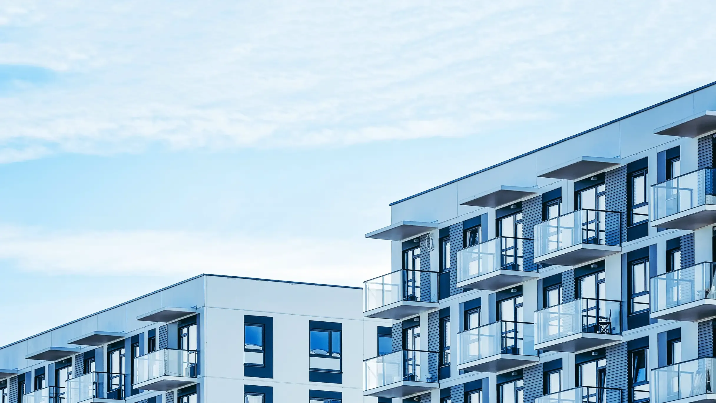 Modern apartment buildings with white facades and glass-fronted balconies under a bright blue sky.
